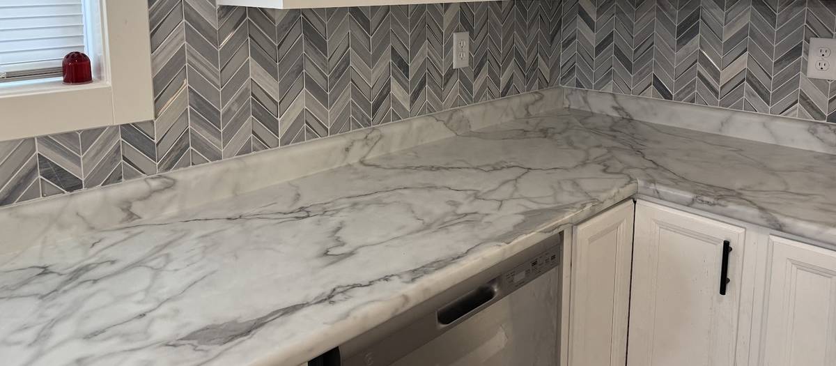 A marble-look kitchen countertop meeting a gray herringbone tile backsplash beneath white cabinets.